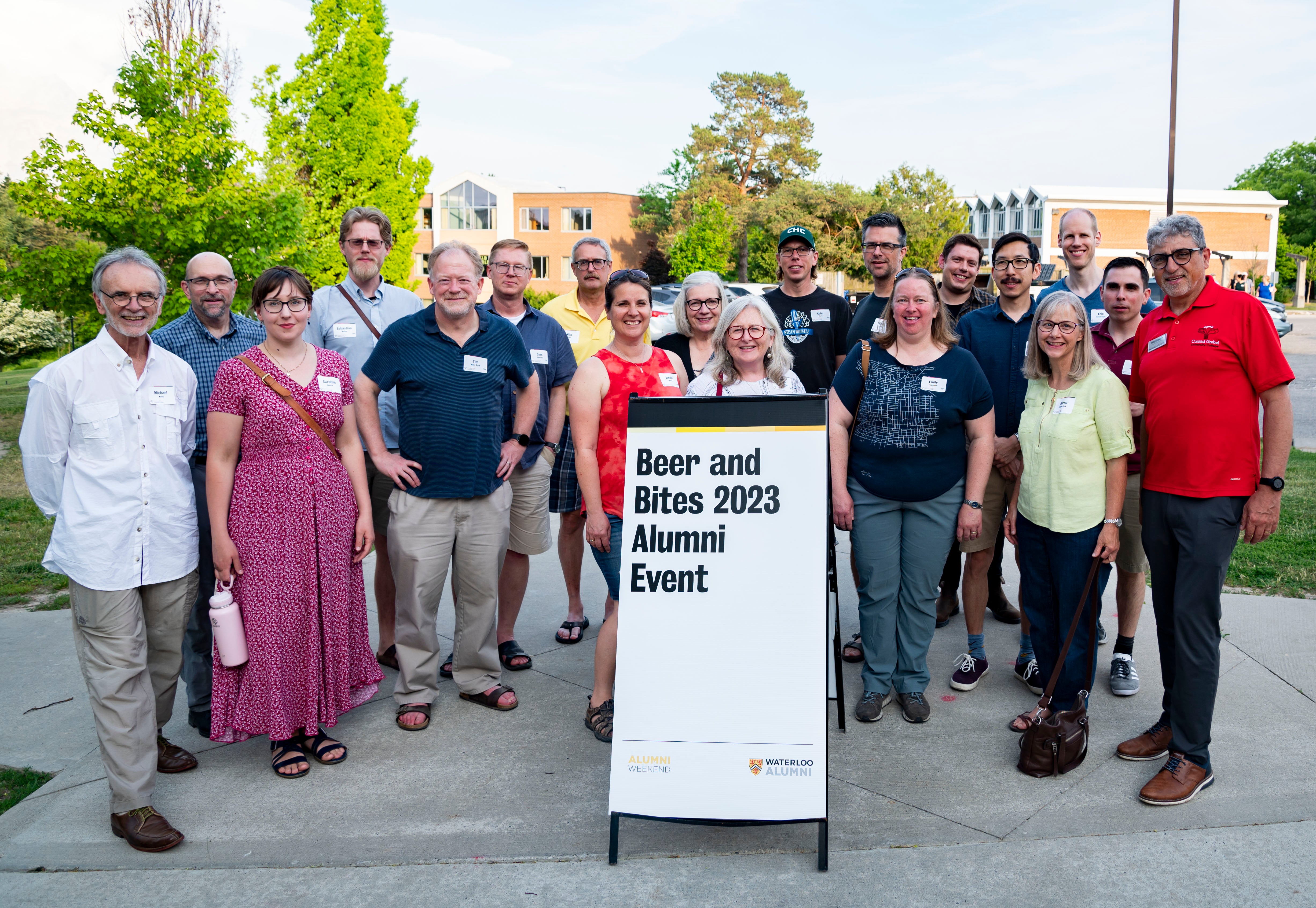 A group picture of twenty alumni outside the beers and bites alumni event sign on a bright sunny evening.