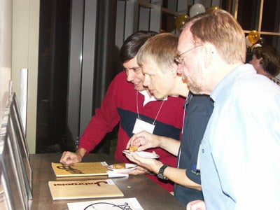 Three men looking at five framed photos that are sitting on a table.