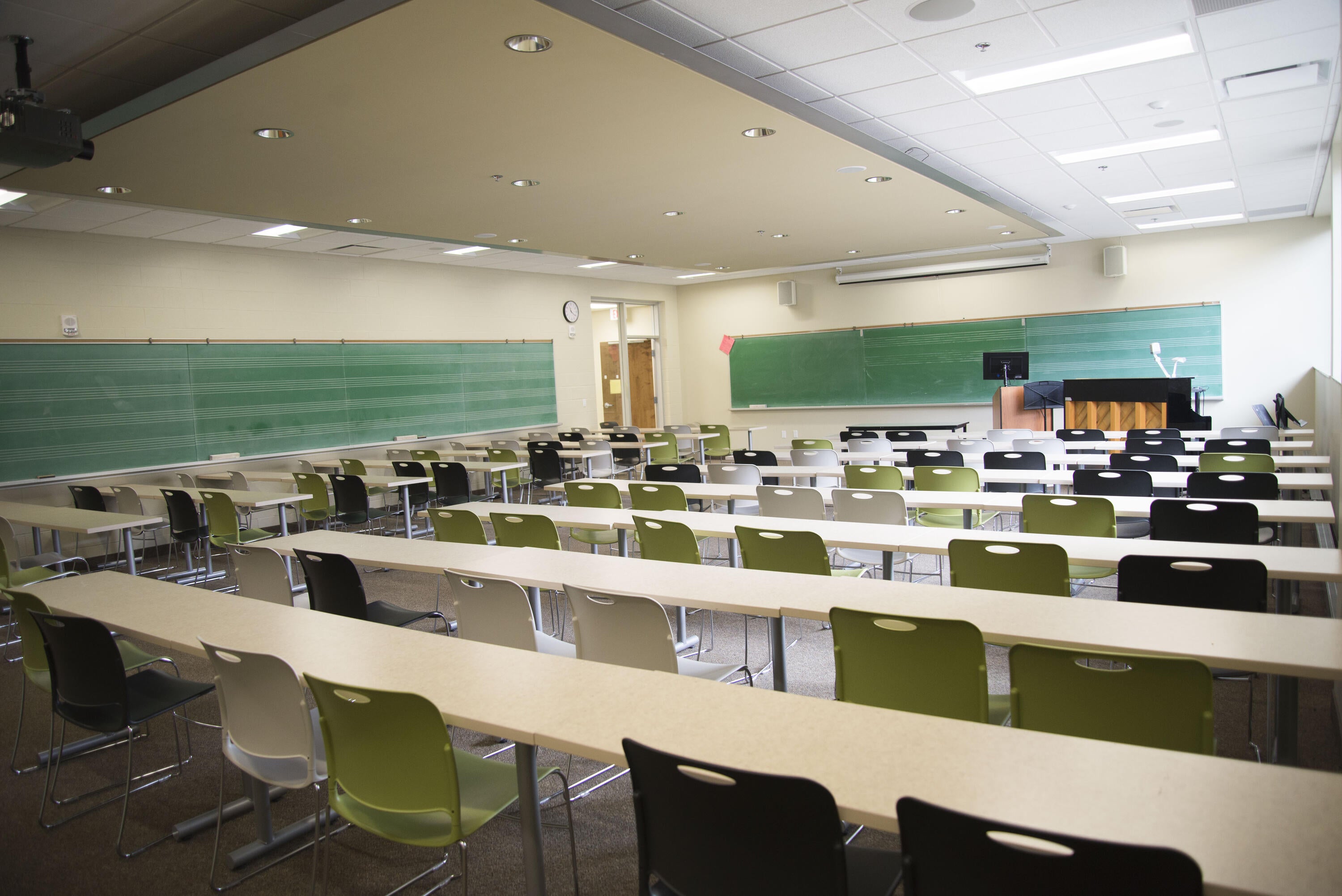Classroom with long tables, chairs, chalkboards, a projector, and an upright piano