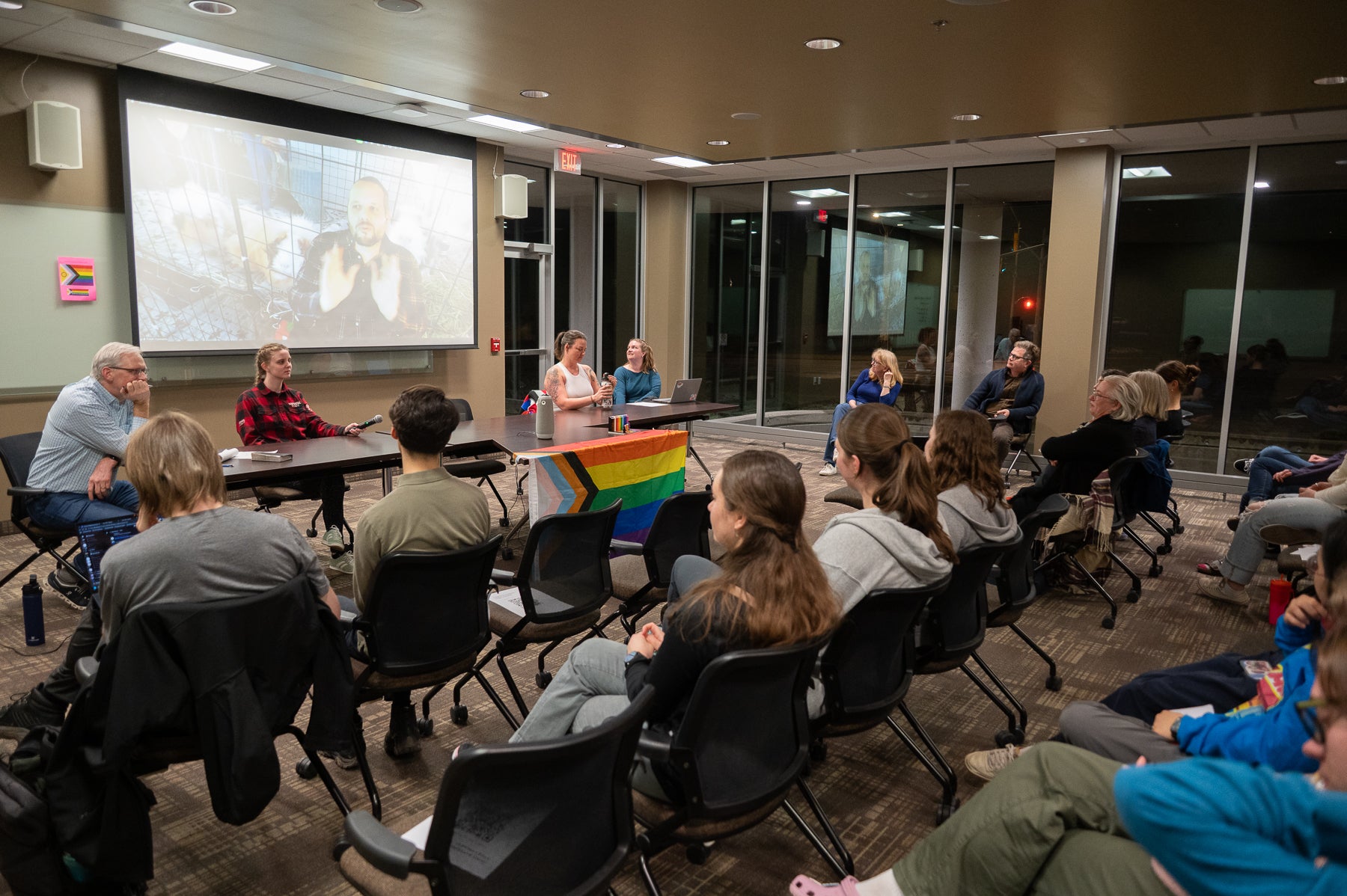 four people sitting at a desk in fornt of a room with a projector screen and a crowd sitting in front of the table.