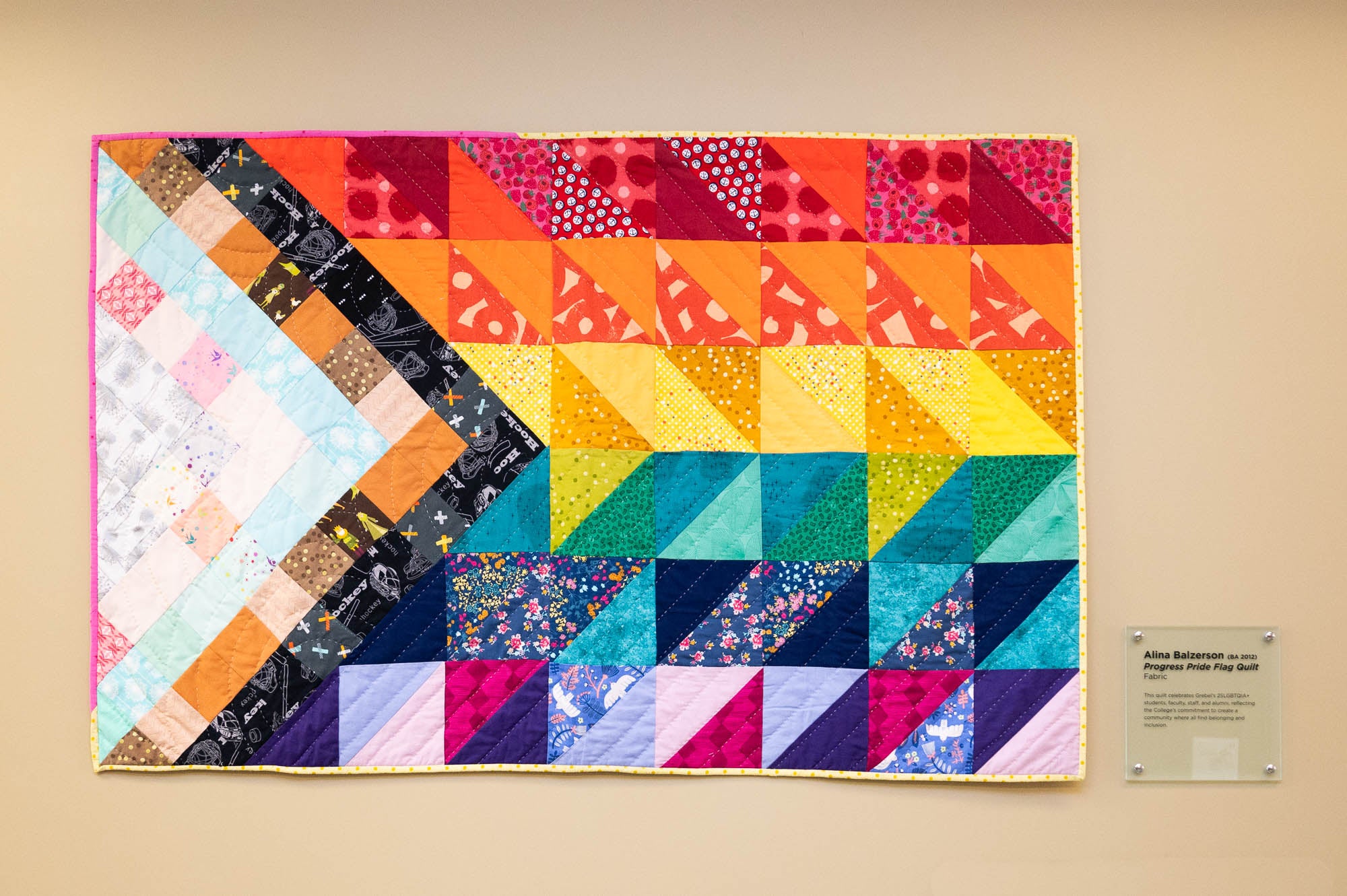 A photograph of a quilt hanging on a wall. The quilted, colourful fabric is made up of triangle shapes to remsemble the pattern of the progress pride flag.