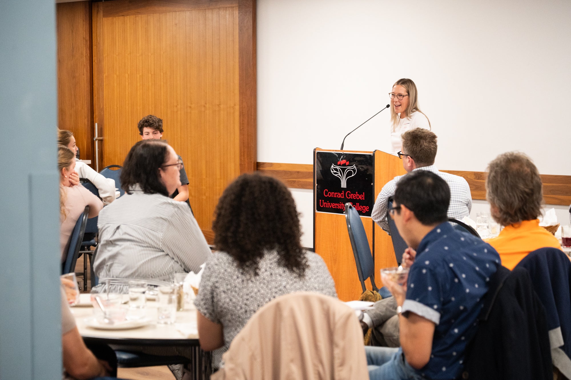 A Grebel alumni speaks at a podium at the front of the dining room