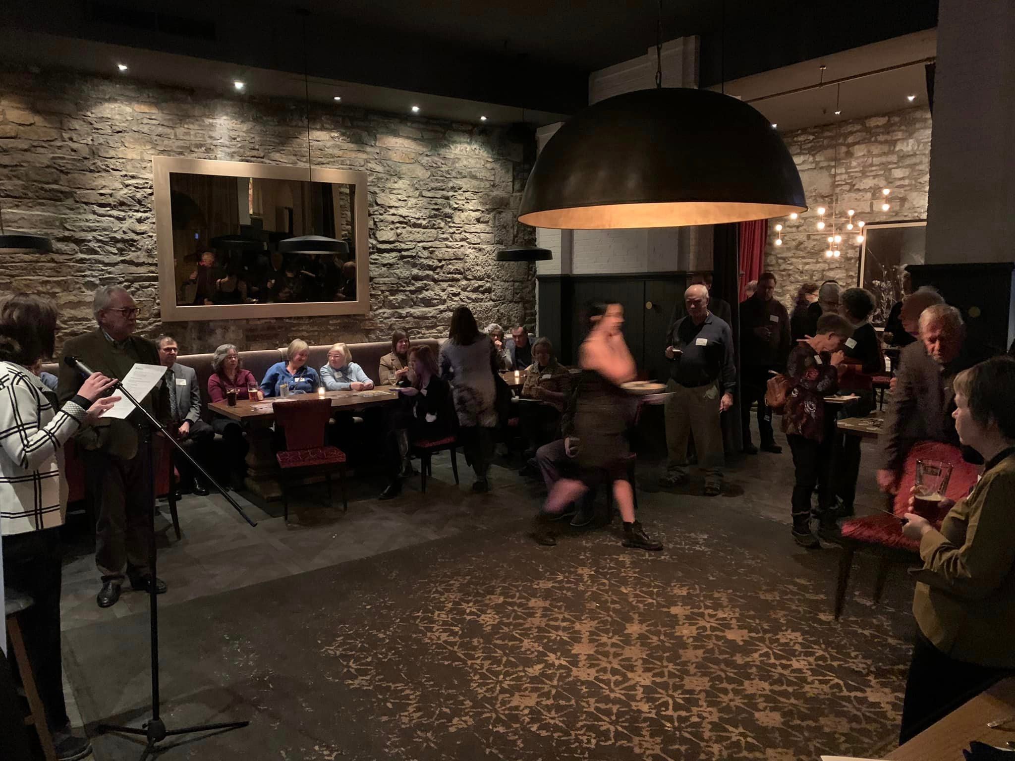A view of the resteraunt room. It's low lit with stone walls, and a cozy vibe. The resteraunt tables along the walls are filled with alumni in conversation.