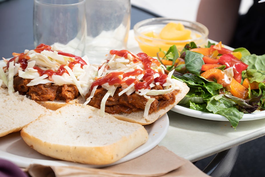 Two pulled pork sandwhiches with coleslaw and siracha sit on a plate next to a lush salad with bell spinach, bell pepper and vinegrette dressing.