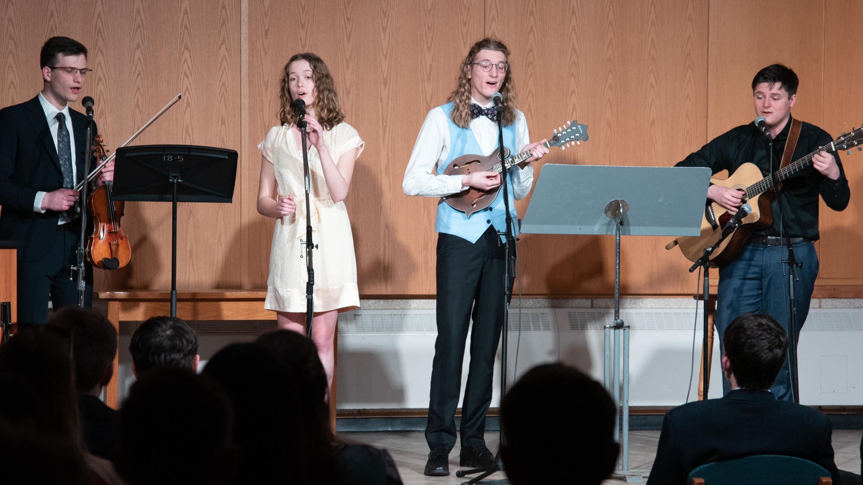 four students singing and playing instruments in front of a crowd 