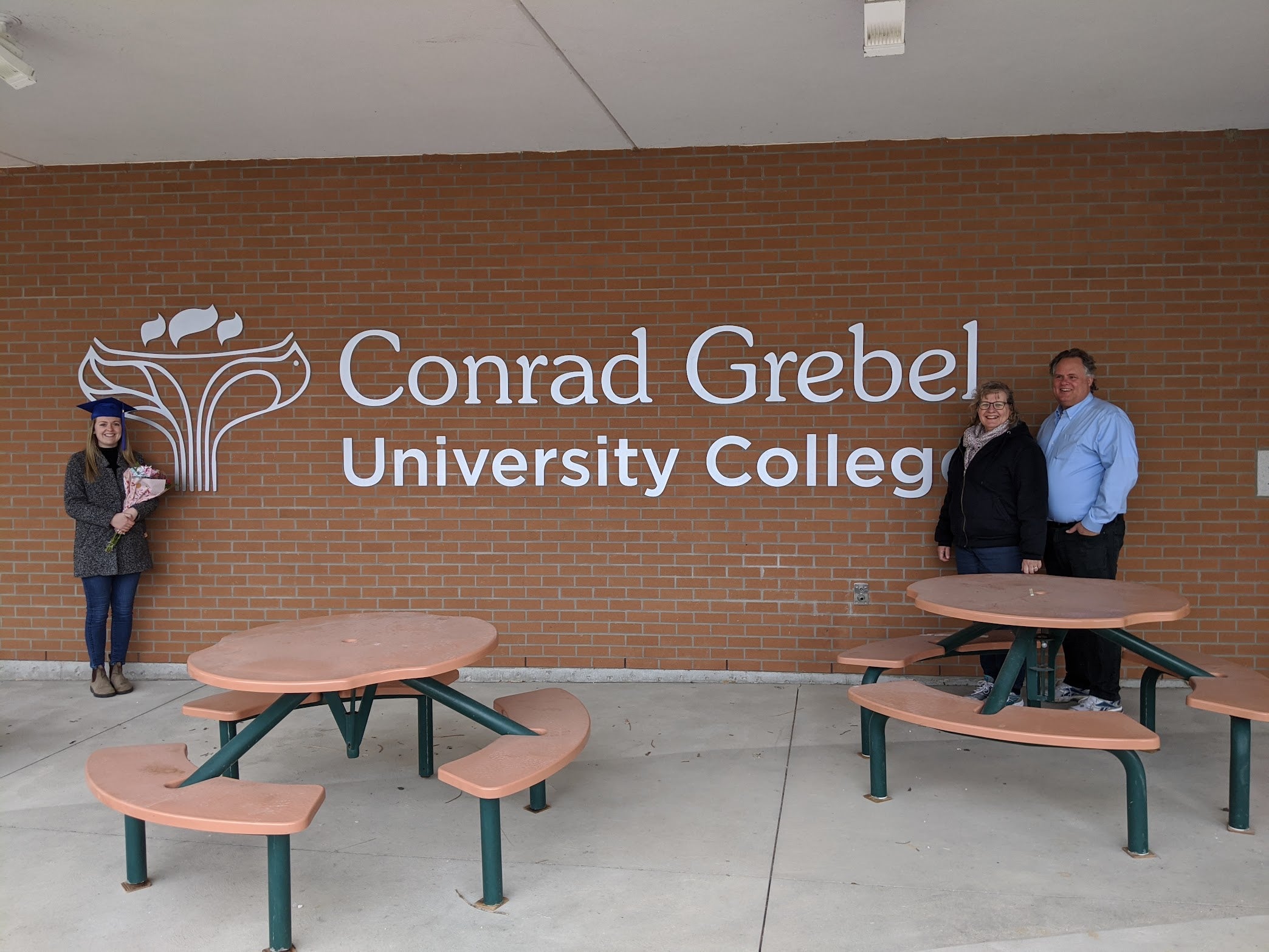 Hannah, wearing a grad cap, stands to the right of the Grebel sign, with Paul and Dolores standing on left, smiling for the camera