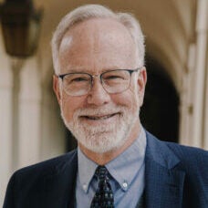 A photo of Mark, wearing a blue suit, smiling. He wear glasses, and has a short white beard.