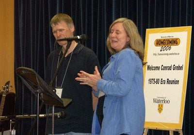 A women speaking to the crowd, with the assistance of a microphone, while a man waits in the background
