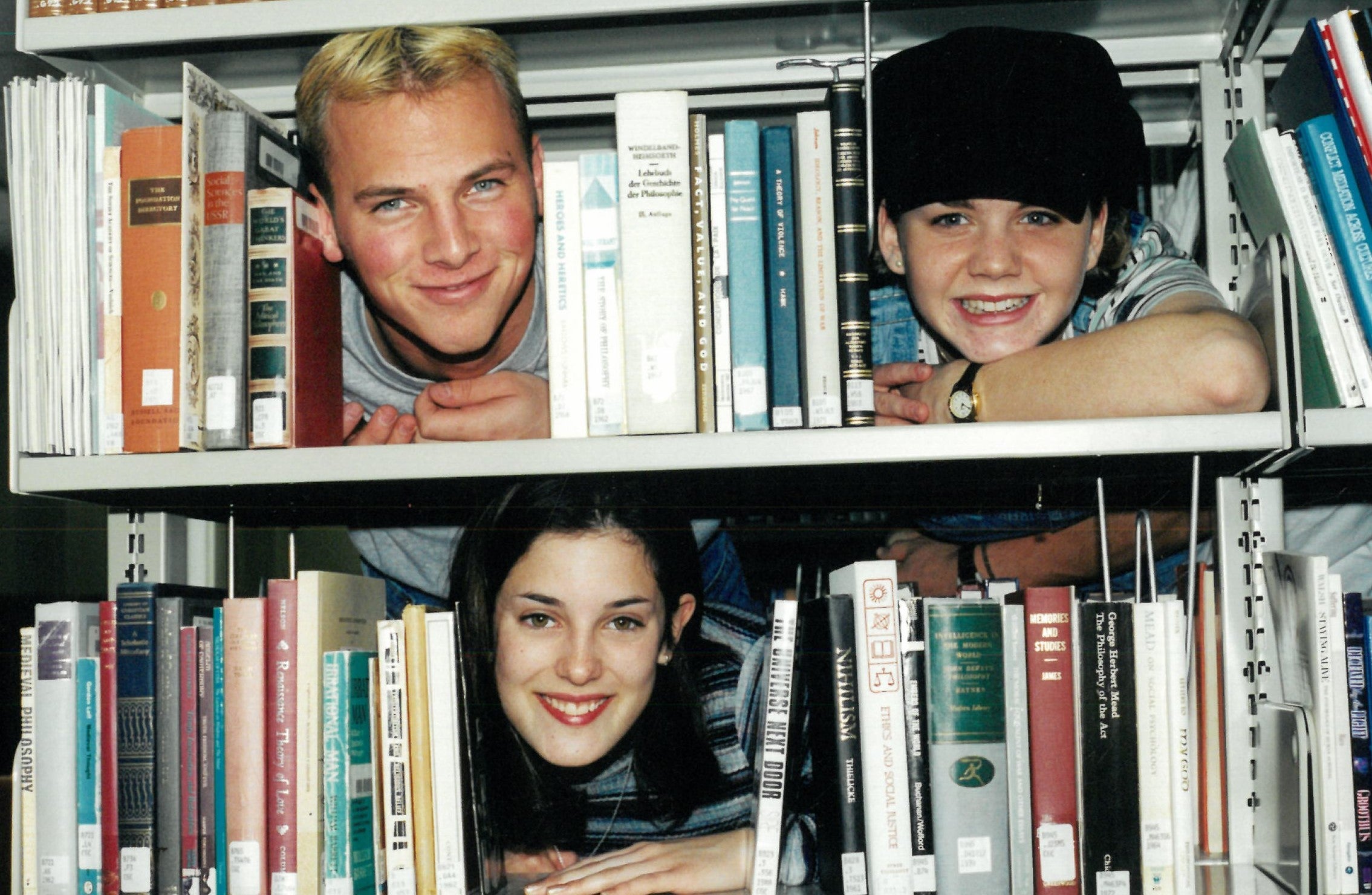 Three students sticking their heads out of bookshelves