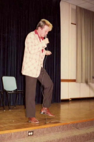 a student stands at a microphone, performing some sort of story (80s)