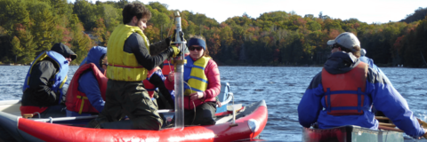 People in a canoe and boat watch as a student uses a sediment coring device off of an inflatable raft in a lake.