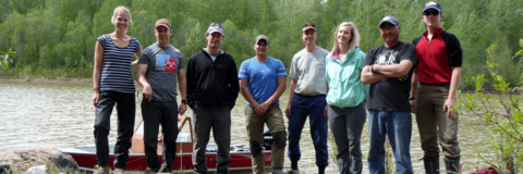 Six men and two women stand on a bedrock outcrop beside a river, in front of a boat.