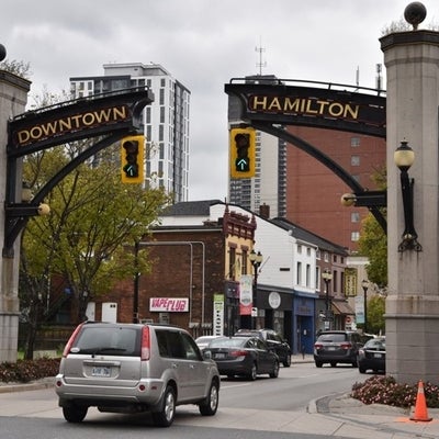 Downtown Hamilton Entry Gate 2 stone columns with black iron grilled arch with downtown Hamilton spelled in gold letters cloudy day with car passing under arch