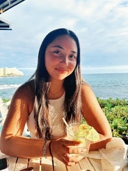 Woman with long black hair and a drink smiling. Trees and beach are in the background.
