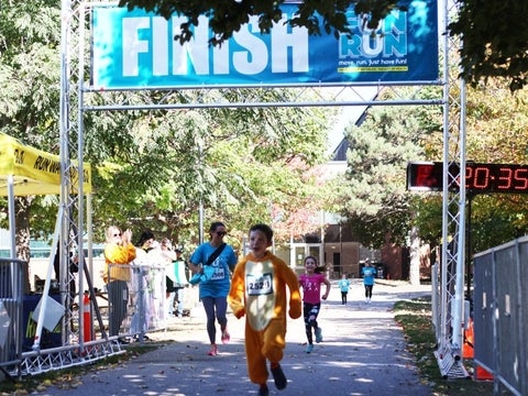 Child in costume crosses the Fun Run finish line with crowd cheering.