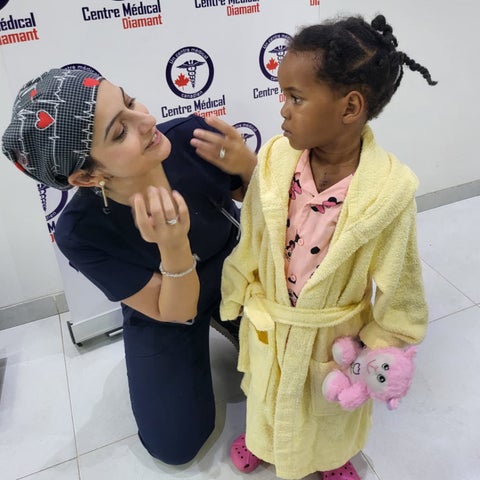 Nurin wears surgical scrubs and kneels smiling beside toddler patient.