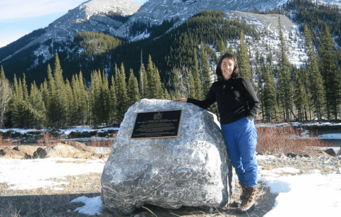 Dawn Carr stands next to large boulder with mountains in background.