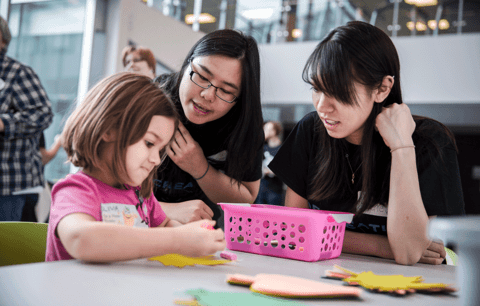 Two students assist young girl drawing at community event.