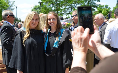 Parent takes photo of two smiling graduates wearing convocation robes.