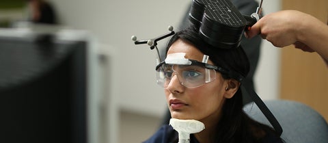 Research subject watches computer monitor while researcher holds TMS device near top of head.