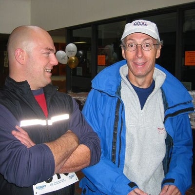Man on right wearing a white hat and blue jacket looking surprised and man on left wearing a black vest smiling