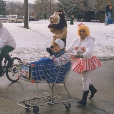 A man dressed up as a woman pushing a shopping cart which a woman is sitting on