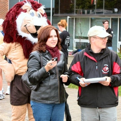 A lion mascot, a woman, and a man standing on the side of the road timing the runners