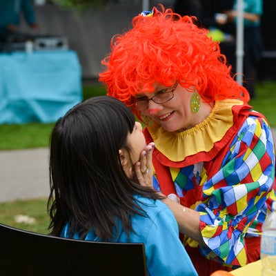 A girl getting her face painted by a woman in a clown suit.