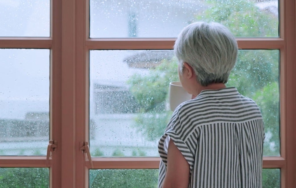 An older adult at home drinking from a cup while looking out a window.