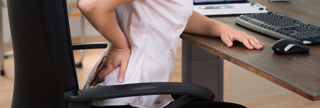 A person sitting at a desk in an office chair holds their lower back, indicating pain.