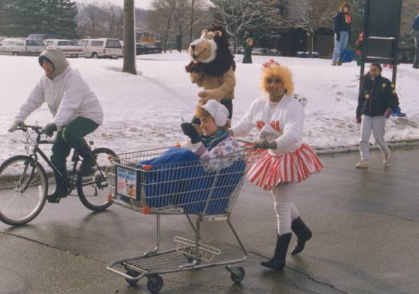 A man dressed up as a woman pushing a shopping cart which a woman is sitting on