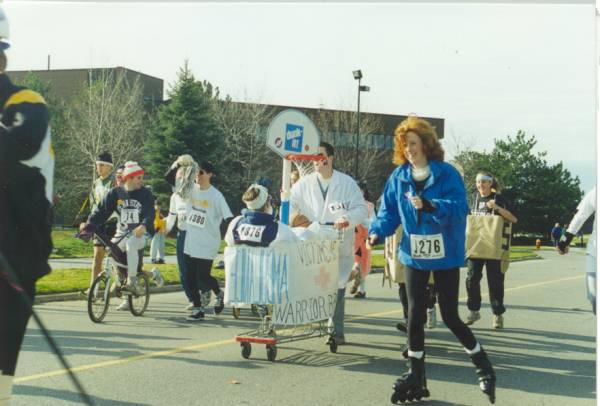 people riding a bike or rollarskating with focus on a participant being pushed by another participant in a shopping cart.