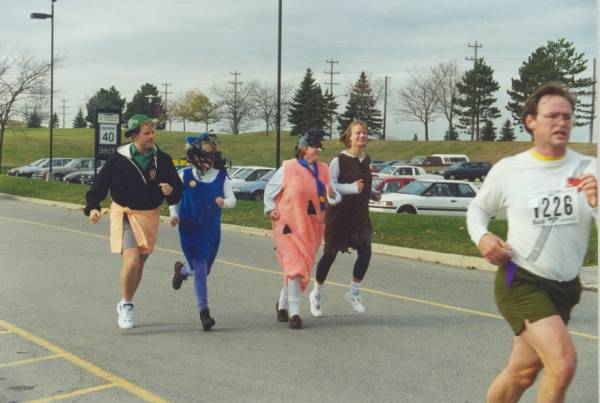 One man running in the front with no costume and other four runners at the back with costumes.