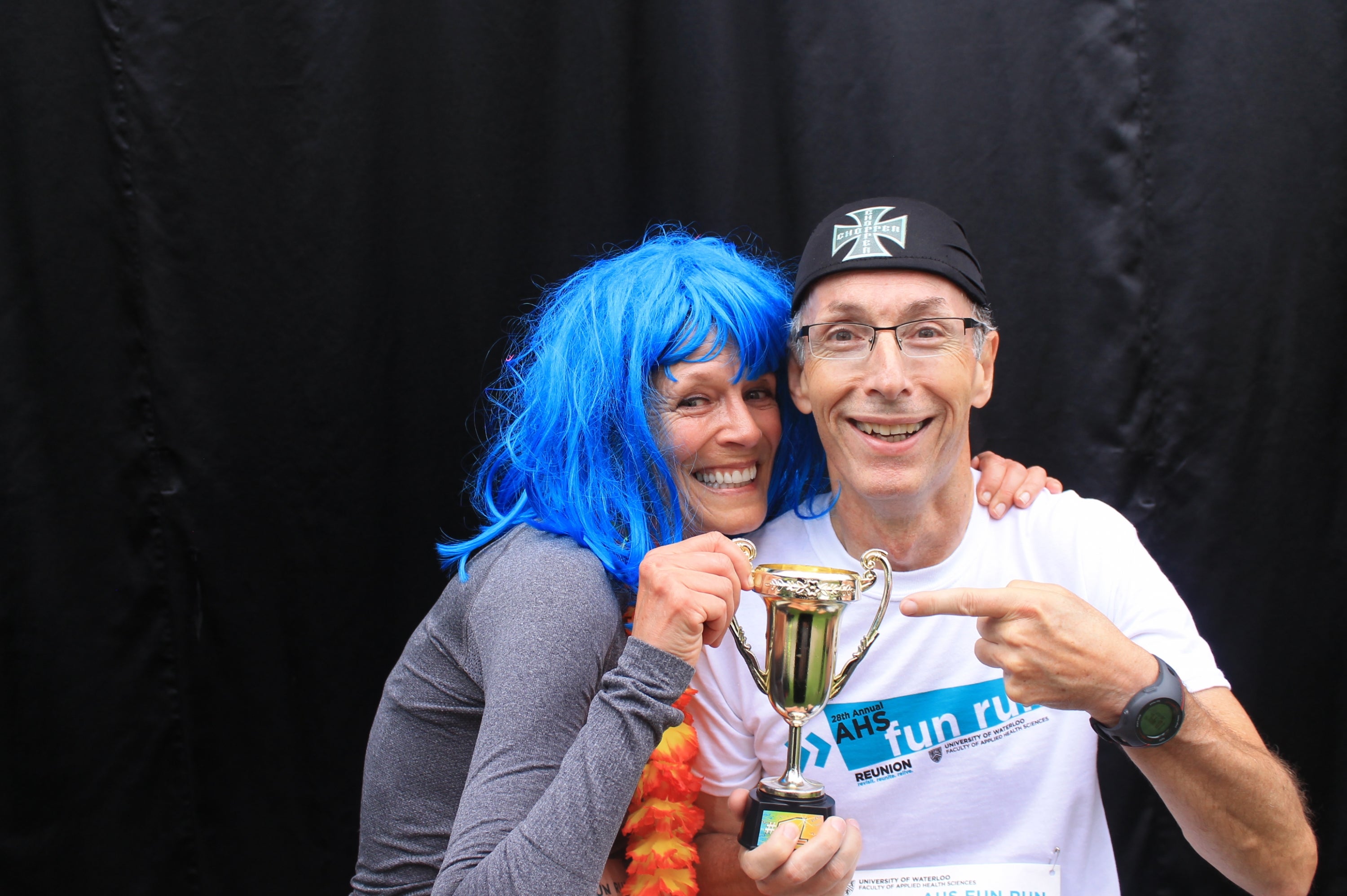 Woman wearing a blue wig and a man with a hat holding a little trophy together.