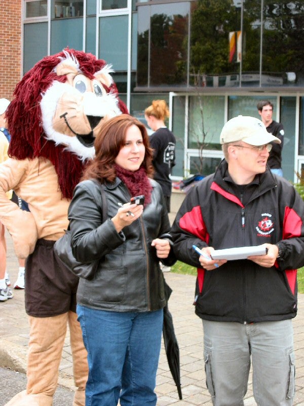 A lion mascot, a woman, and a man standing on the side of the road timing the runners
