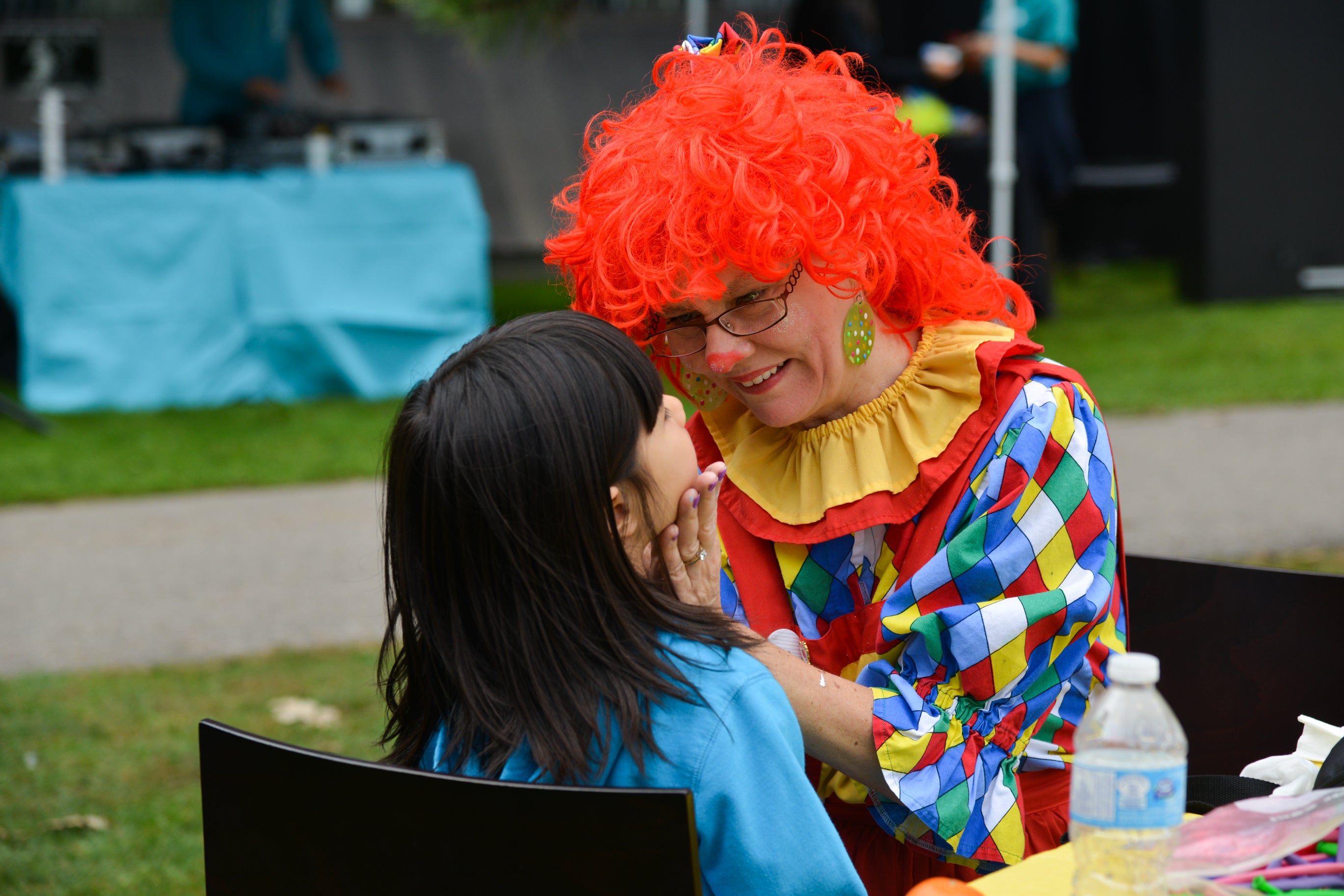 A girl getting her face painted by a woman in a clown suit.