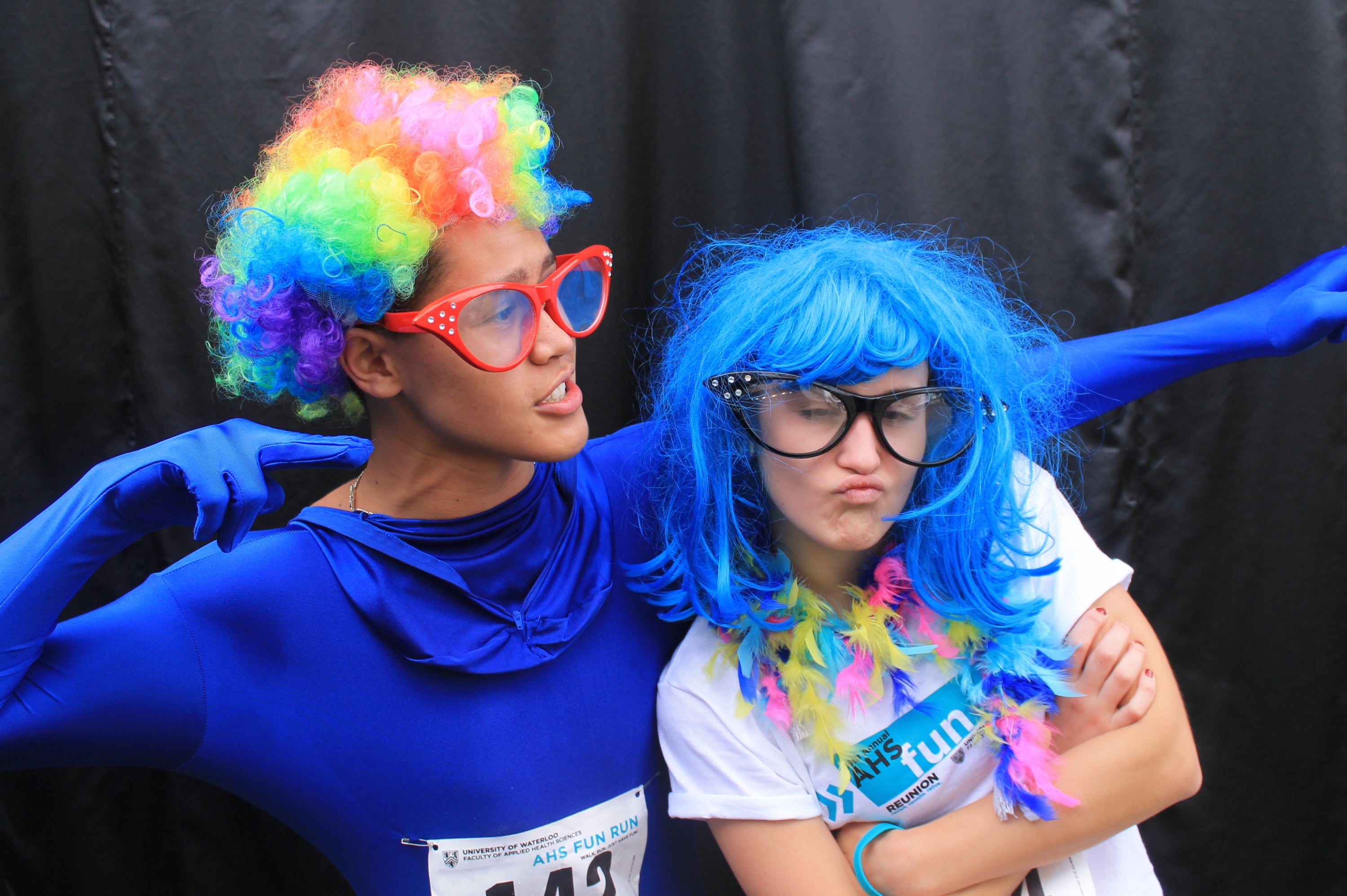 A man wearing a full blue body suit, overzised glasses and a wig while a woman is crossing her arms with glasses and a blue wig.