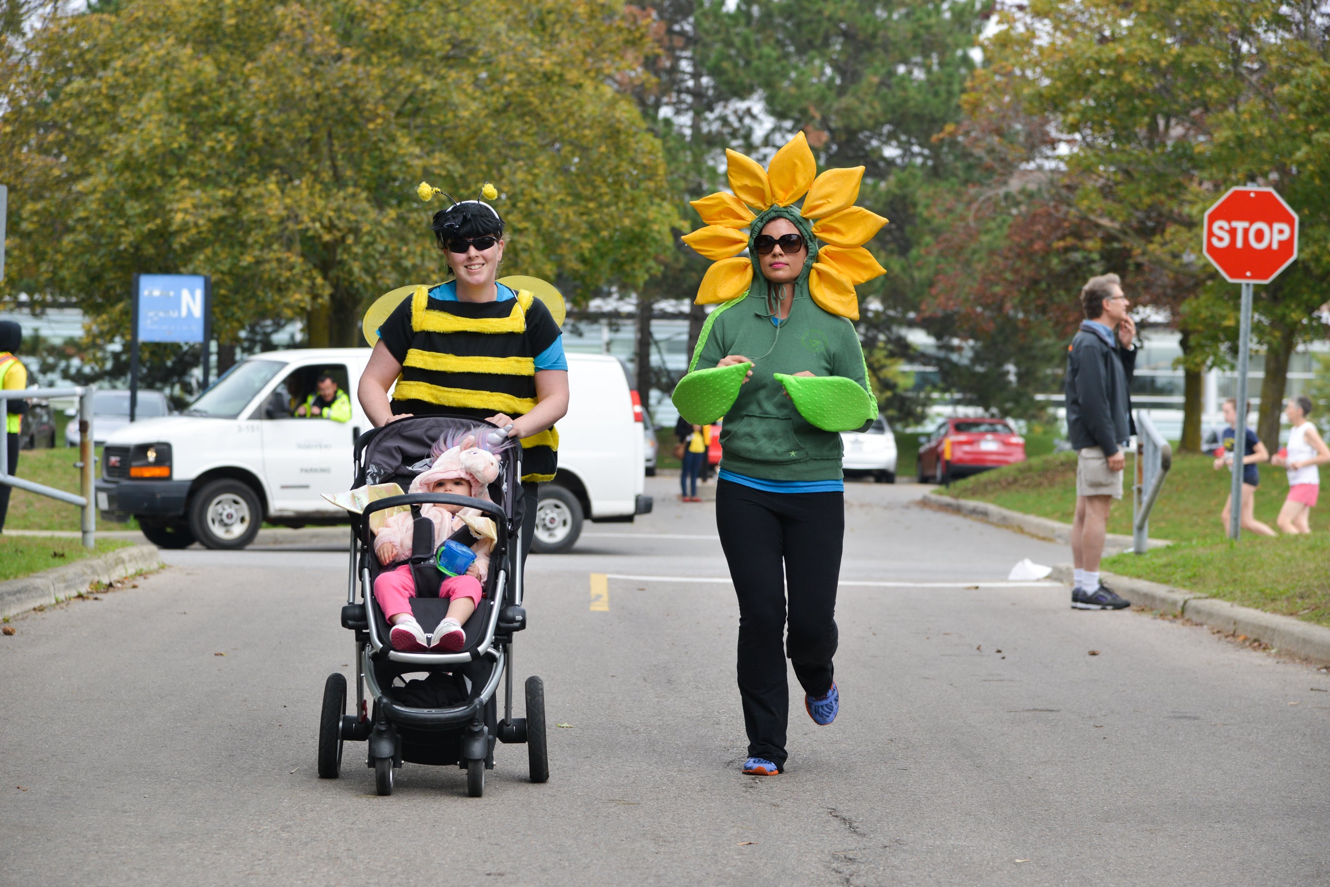 Lady in a bee suit pushing her daughter in a carriage while another lady dressed as a flowe runs beside her.