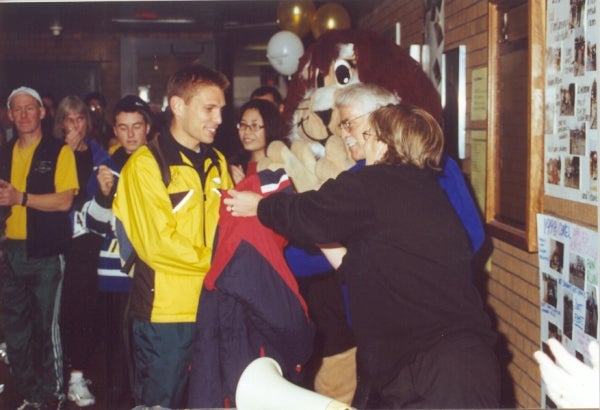 Gathering of people with two elder individuals beside a bear mascot handing a jacket to a young male