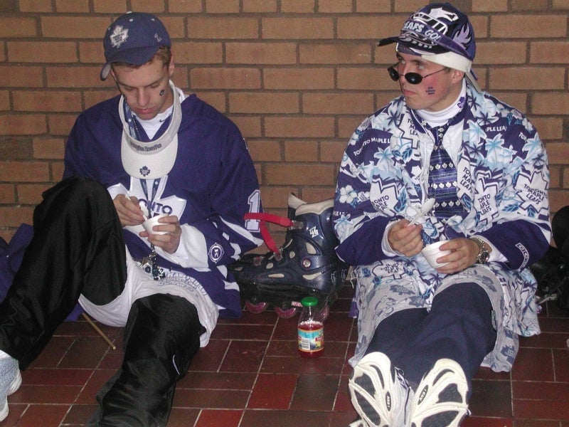Two male individuals sitting on the ground wearing Toronto Maple Leafs hockey team fan gear.