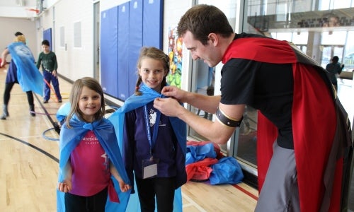 University student wearing red cape ties super hero capes on young visitors.