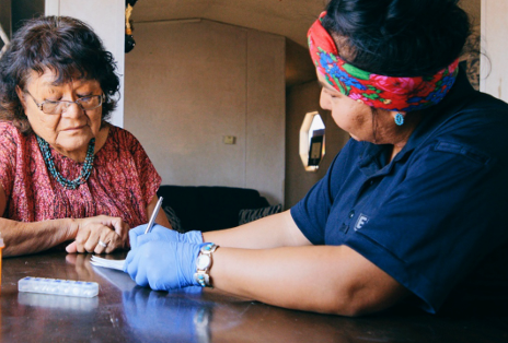 An Indigenous medical professional sits a table with an elderly woman.