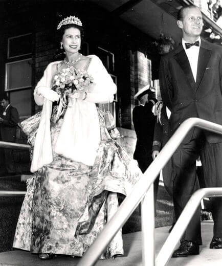 A black and white image from 1959 of the Queen and Prince Phillip visiting Stratford, walking down steps with the queen holding flowers and the prince in a tuxedo