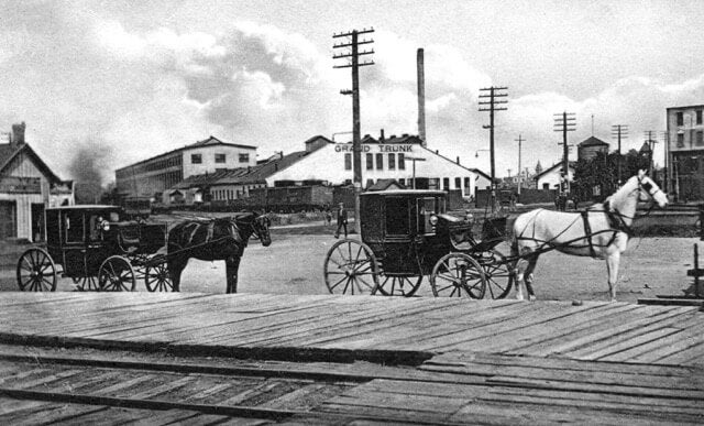 A historic black and white photograph of a viewpoint of the Stratfor shops in the background with two horses carrying buggies in front