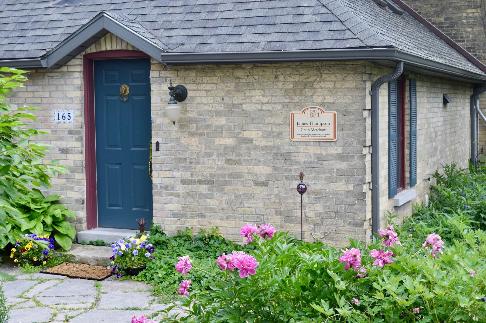 The corner of a stone home with a plaque on the wall and garden beside, clear to read by pedestrians
