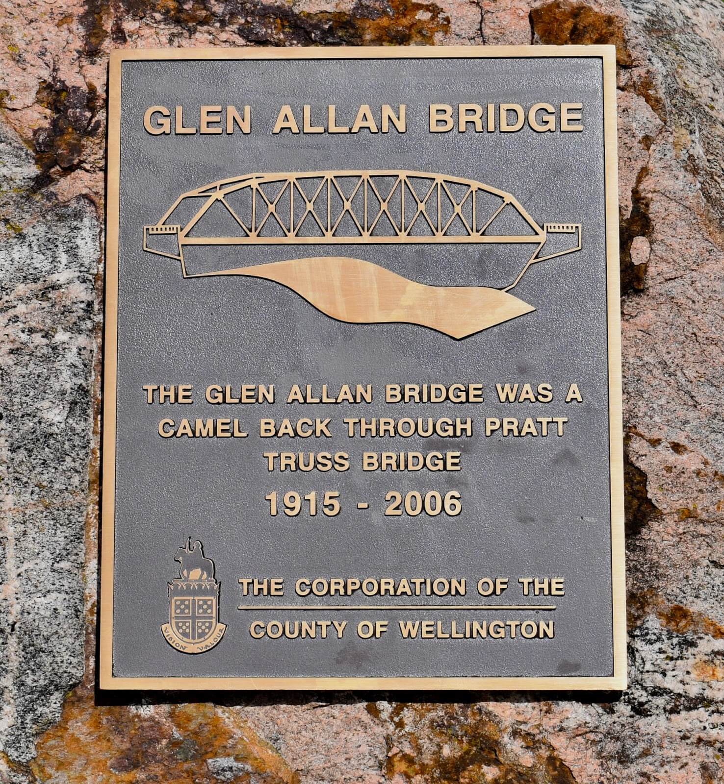 A metal plaque for a ruined bridge in Glen Allan, Ontario reading: "The Glen Allan Bridge was a camel back through pratt truss bridge 1915-2006, The Corporation of the County of Wellington"