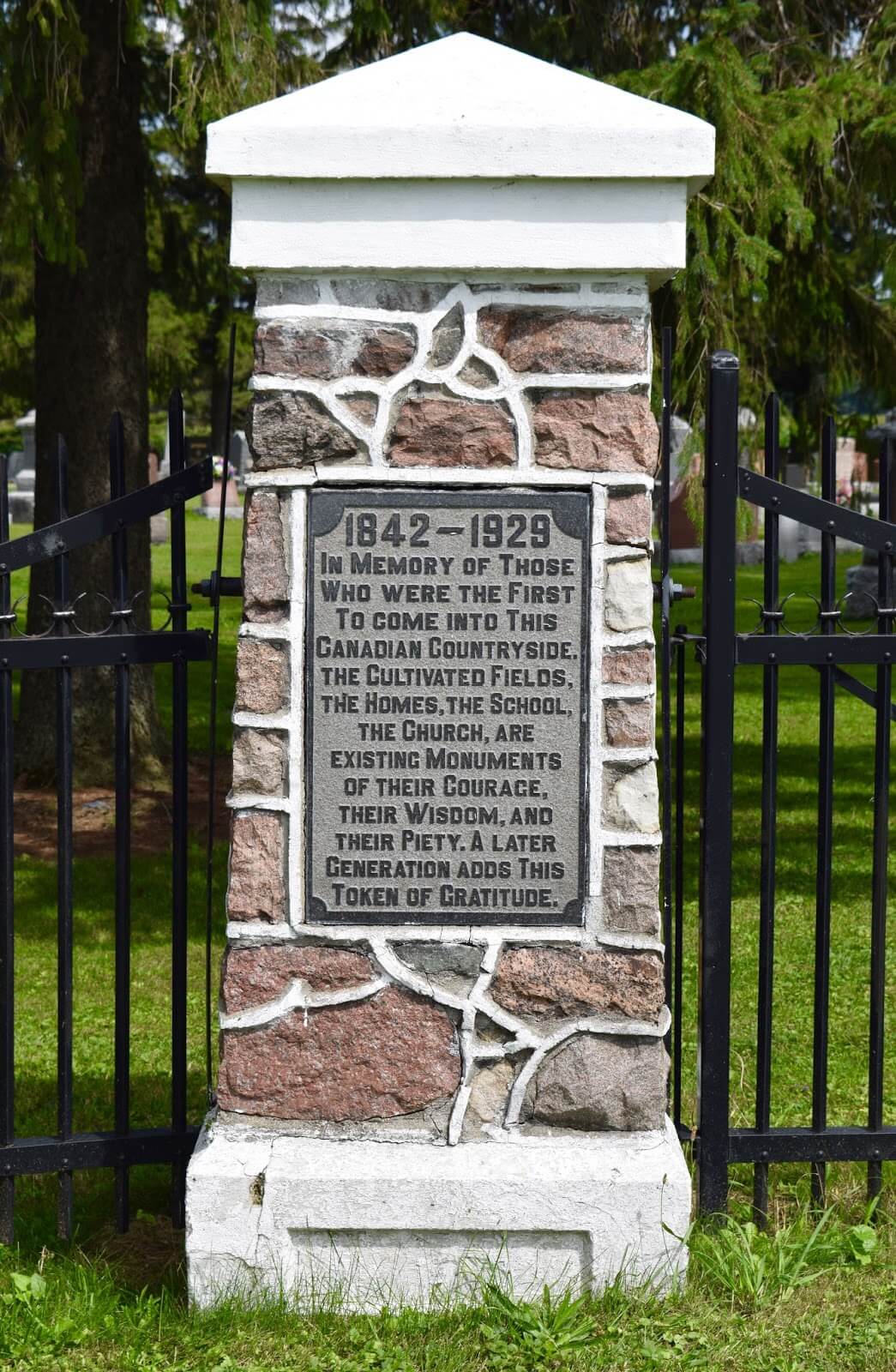A plaque stating 1842-1929, In memory of those who were the first to come into this Canadian countryside. The cultivated fields, the homes, the school, the church, are existing monuments of their courage, their wisdom, and their piety. A later generation adds this token of gratitude.