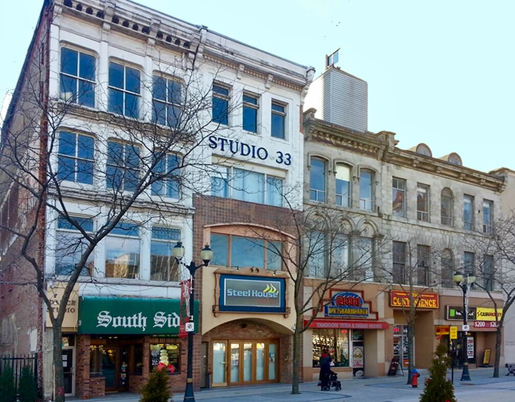 The existing facade of Gore Street, Hamilton, Ontario as it is today, brick and mortar frontages with pedestrians and trees in front