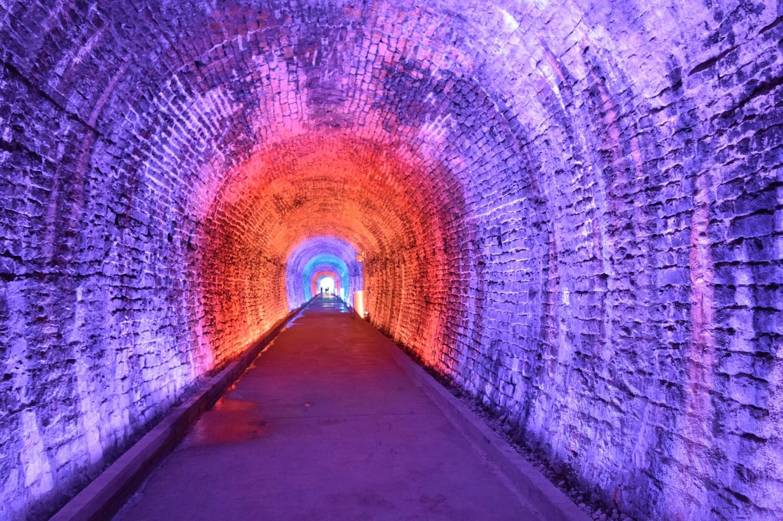 A photograph of an old railway tunnel used as a public art project with red and blue lights highlighting the interior of the tunnel