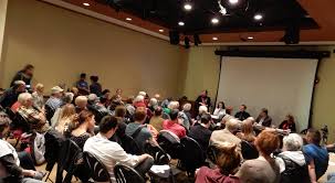 A photograph of a crowded conference room from behind, rows of people sitting in front of a blank projector screen